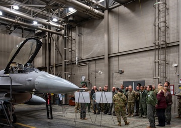 A group of approximately 20-25 military and civilian personnel are gathered inside a large aircraft maintenance hangar for a briefing or presentation. They are standing in front of two easel-mounted whiteboards/flip charts near an F-16 Fighting Falcon fighter jet with tail markings 'AK' and number '554'. The setting appears to be a U.S. Air Force facility, likely in Alaska based on the 'AK' tail code.