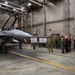 A group of approximately 20-25 military and civilian personnel are gathered inside a large aircraft maintenance hangar for a briefing or presentation. They are standing in front of two easel-mounted whiteboards/flip charts near an F-16 Fighting Falcon fighter jet with tail markings 'AK' and number '554'. The setting appears to be a U.S. Air Force facility, likely in Alaska based on the 'AK' tail code.