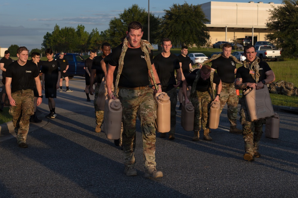 A group of military personnel are conducting a physical training exercise on a paved road at what appears to be a military installation during early morning or late afternoon light. Several individuals in the foreground are wearing camouflage uniforms and carrying large military-style water or fuel jerry cans in each hand. Others in the background are wearing black PT (physical training) shirts with unit markings and camouflage pants.