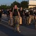 A group of military personnel are conducting a physical training exercise on a paved road at what appears to be a military installation during early morning or late afternoon light. Several individuals in the foreground are wearing camouflage uniforms and carrying large military-style water or fuel jerry cans in each hand. Others in the background are wearing black PT (physical training) shirts with unit markings and camouflage pants.