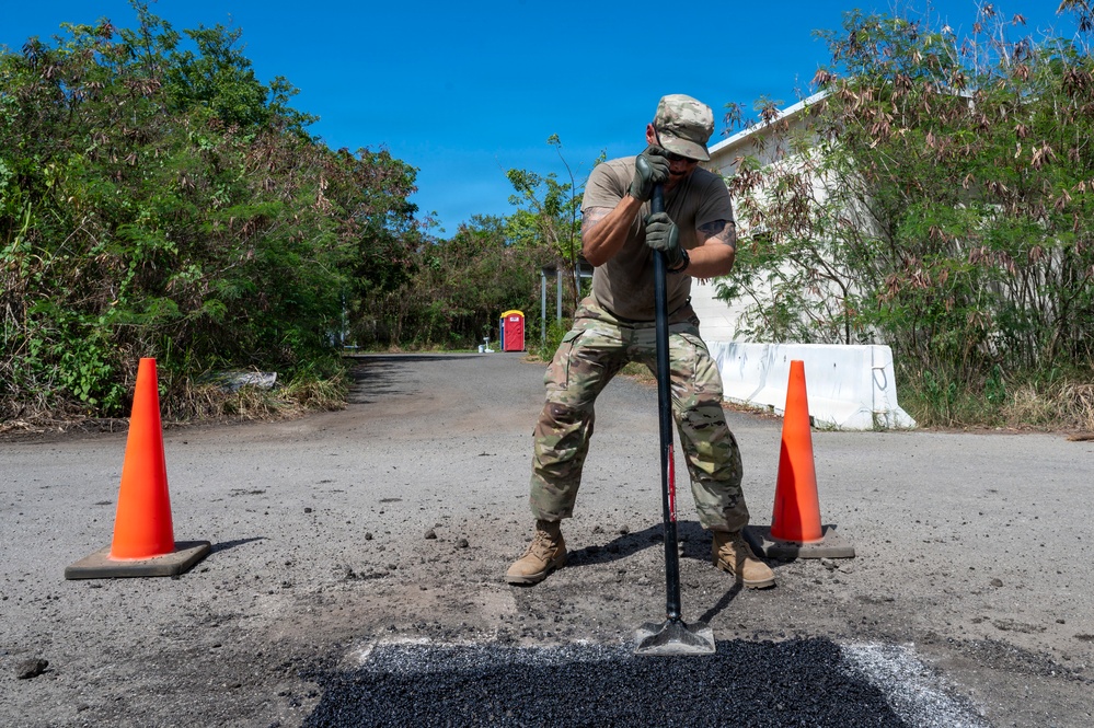 A military service member in camouflage uniform is performing road repair work, using a tamping tool to compact fresh asphalt into a pothole. The work area is marked with two orange traffic safety cones. The setting appears to be a damaged road area, possibly following a natural disaster, with storm-damaged vegetation visible in the background.