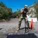 A military service member in camouflage uniform is performing road repair work, using a tamping tool to compact fresh asphalt into a pothole. The work area is marked with two orange traffic safety cones. The setting appears to be a damaged road area, possibly following a natural disaster, with storm-damaged vegetation visible in the background.