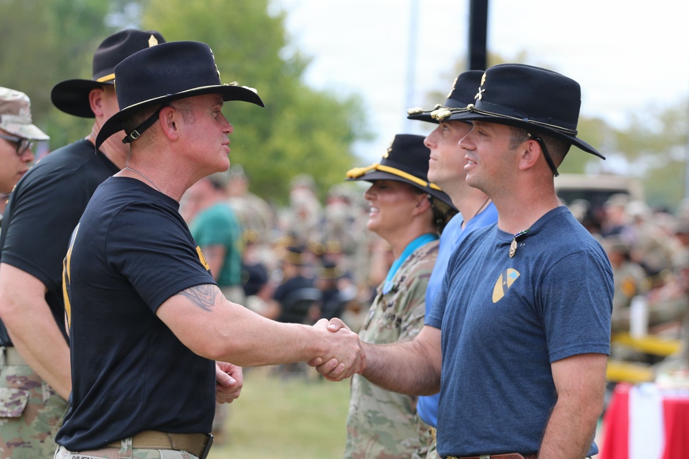 Two men in U.S. Army cavalry-style black Stetsons and PT (physical training) uniforms are shaking hands at what appears to be a military ceremony or event. Both wear black t-shirts with gold unit insignia, consistent with 1st Cavalry Division markings. A crowd of soldiers and attendees is visible in the background.