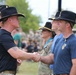 Two men in U.S. Army cavalry-style black Stetsons and PT (physical training) uniforms are shaking hands at what appears to be a military ceremony or event. Both wear black t-shirts with gold unit insignia, consistent with 1st Cavalry Division markings. A crowd of soldiers and attendees is visible in the background.
