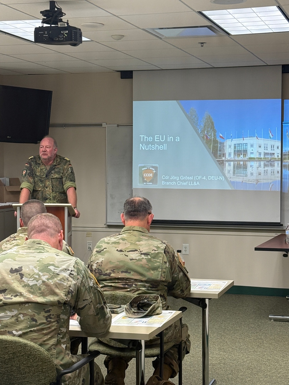 A military briefing is taking place in a classroom setting where a German Bundeswehr officer is presenting a slide titled 'The EU in a Nutshell' to an audience of soldiers in camouflage uniforms. The presenter is identified on the slide as Cdr Jörg Grössl (OF-4, DEU-N), Branch Chief LL&amp;A, affiliated with CCOE (Civil-Military Cooperation Centre of Excellence). The audience members, wearing what appears to be US Army MultiCam/OCP uniforms, are seated at desks with printed materials in front of them.