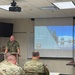 A military briefing is taking place in a classroom setting where a German Bundeswehr officer is presenting a slide titled 'The EU in a Nutshell' to an audience of soldiers in camouflage uniforms. The presenter is identified on the slide as Cdr Jörg Grössl (OF-4, DEU-N), Branch Chief LL&amp;A, affiliated with CCOE (Civil-Military Cooperation Centre of Excellence). The audience members, wearing what appears to be US Army MultiCam/OCP uniforms, are seated at desks with printed materials in front of them.