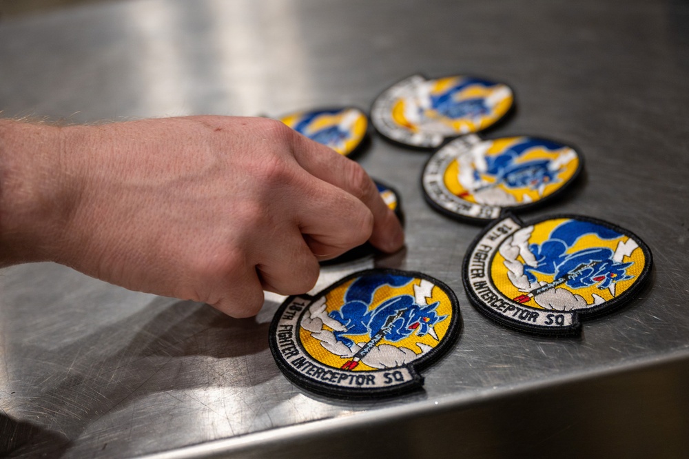 A hand is arranging multiple embroidered military patches on a stainless steel surface. The patches belong to the '18th Fighter Interceptor Squadron' and feature a blue eagle or bird design on a yellow and white background with a black border. Multiple identical patches are visible, suggesting inventory management or distribution activity.