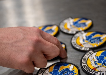 A hand is arranging multiple embroidered military patches on a stainless steel surface. The patches belong to the '18th Fighter Interceptor Squadron' and feature a blue eagle or bird design on a yellow and white background with a black border. Multiple identical patches are visible, suggesting inventory management or distribution activity.