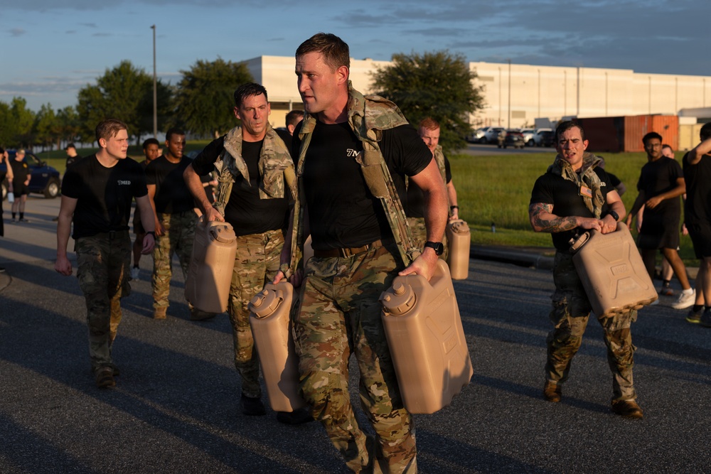 A group of military personnel in camouflage uniforms and black PT (physical training) shirts are conducting a physical training exercise carrying tan/beige military jerry cans (water or fuel containers). The exercise appears to take place on a paved area near a military installation or base, photographed during early morning or late afternoon golden hour lighting. Multiple participants are visible carrying the heavy containers in what appears to be a structured fitness or endurance training event.