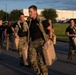 A group of military personnel in camouflage uniforms and black PT (physical training) shirts are conducting a physical training exercise carrying tan/beige military jerry cans (water or fuel containers). The exercise appears to take place on a paved area near a military installation or base, photographed during early morning or late afternoon golden hour lighting. Multiple participants are visible carrying the heavy containers in what appears to be a structured fitness or endurance training event.