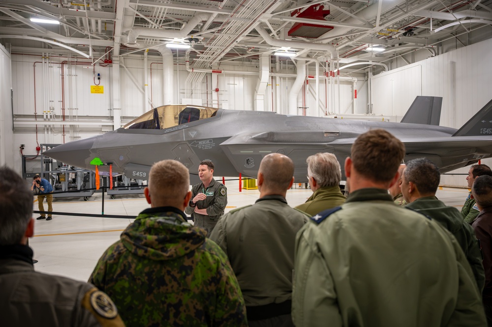A military briefing is taking place inside an aircraft hangar featuring at least one F-35 Lightning II stealth fighter jet with tail number 5459 visible. A uniformed individual in a green flight suit appears to be presenting or briefing a group of mixed military personnel wearing various uniforms, including camouflage and olive flight suits. A photographer is visible in the background documenting the event.