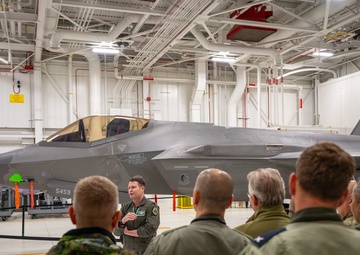 A military briefing is taking place inside an aircraft hangar featuring at least one F-35 Lightning II stealth fighter jet with tail number 5459 visible. A uniformed individual in a green flight suit appears to be presenting or briefing a group of mixed military personnel wearing various uniforms, including camouflage and olive flight suits. A photographer is visible in the background documenting the event.