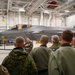A military briefing is taking place inside an aircraft hangar featuring at least one F-35 Lightning II stealth fighter jet with tail number 5459 visible. A uniformed individual in a green flight suit appears to be presenting or briefing a group of mixed military personnel wearing various uniforms, including camouflage and olive flight suits. A photographer is visible in the background documenting the event.