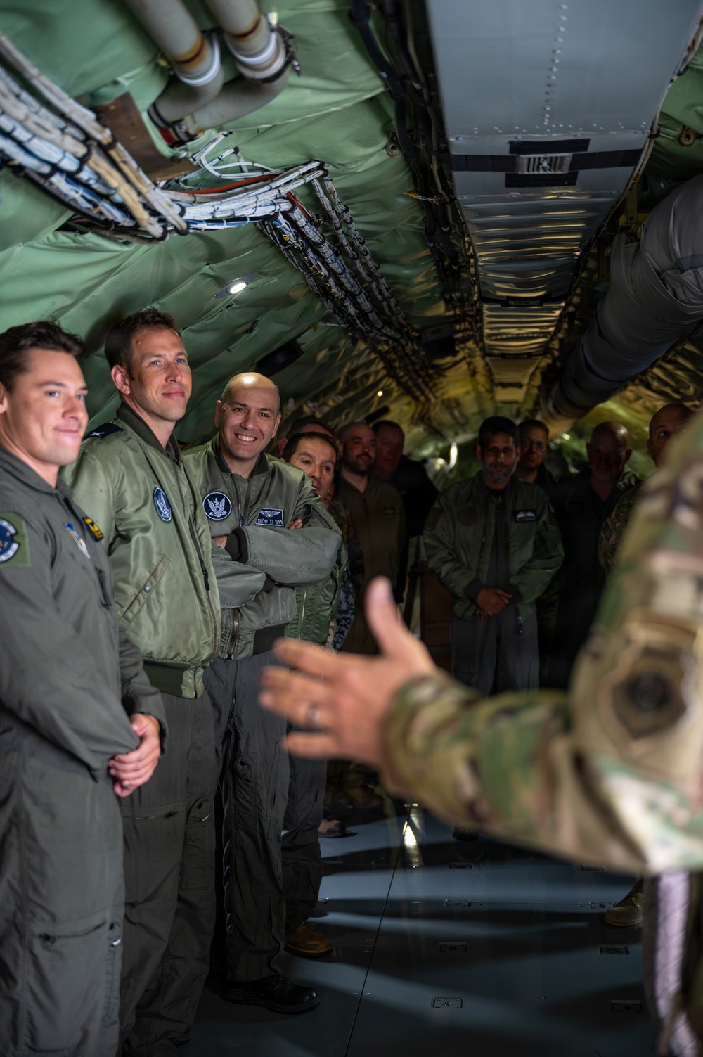 Military personnel in flight suits are gathered inside the interior of a large military aircraft, likely during a briefing or tour. A person in multicam/OCP uniform on the right side appears to be gesturing and speaking to the group. The group includes personnel wearing different national military flight suits, suggesting a multinational or joint military exercise.