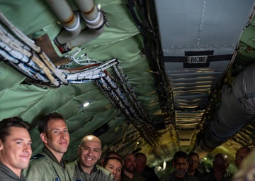 Military personnel in flight suits are gathered inside the interior of a large military aircraft, likely during a briefing or tour. A person in multicam/OCP uniform on the right side appears to be gesturing and speaking to the group. The group includes personnel wearing different national military flight suits, suggesting a multinational or joint military exercise.