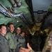Military personnel in flight suits are gathered inside the interior of a large military aircraft, likely during a briefing or tour. A person in multicam/OCP uniform on the right side appears to be gesturing and speaking to the group. The group includes personnel wearing different national military flight suits, suggesting a multinational or joint military exercise.