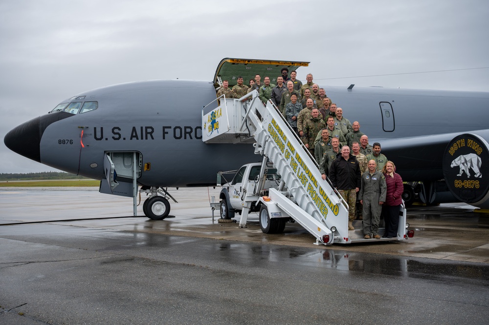 A large group of U.S. Air Force personnel and civilians pose for a group photo on and around a mobile staircase positioned at the door of a U.S. Air Force aircraft, tail number 8876. The aircraft bears the markings of the 168th Wing and the inscription 'Alaska Air National Guard - Guardians of the Last Frontier.' The wet tarmac and overcast sky suggest the photo was taken in Alaska, consistent with the unit's known base at Eielson or Fairbanks.