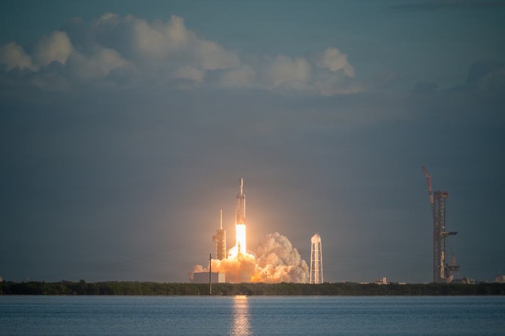 A large rocket is captured mid-launch from a coastal launch facility, producing intense flames and billowing white and orange exhaust clouds at its base. The launch is photographed from across a body of water, with a second launch structure or tower visible to the right in the background. The image appears to depict a SpaceX Falcon Heavy or similar multi-core rocket based on the visible triple-booster configuration.