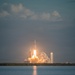 A large rocket is captured mid-launch from a coastal launch facility, producing intense flames and billowing white and orange exhaust clouds at its base. The launch is photographed from across a body of water, with a second launch structure or tower visible to the right in the background. The image appears to depict a SpaceX Falcon Heavy or similar multi-core rocket based on the visible triple-booster configuration.