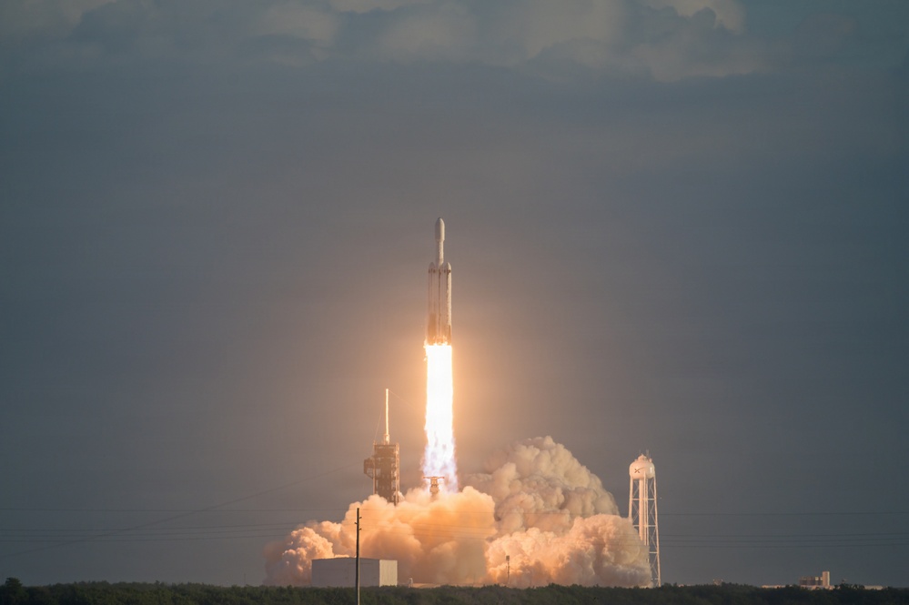 A large rocket is captured mid-launch from what appears to be a coastal launch facility, producing intense flames and billowing clouds of exhaust smoke. The rocket's triple-core configuration is visible, consistent with a heavy-lift launch vehicle. The image was taken during daylight under overcast skies, with the launch pad infrastructure visible in the background.