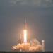 A large rocket is captured mid-launch from what appears to be a coastal launch facility, producing intense flames and billowing clouds of exhaust smoke. The rocket's triple-core configuration is visible, consistent with a heavy-lift launch vehicle. The image was taken during daylight under overcast skies, with the launch pad infrastructure visible in the background.