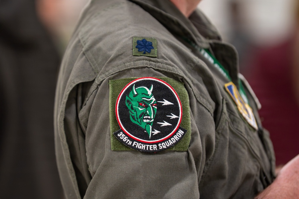 The image shows a close-up of a military flight suit sleeve bearing a squadron patch and rank insignia. The patch identifies the wearer as a member of the 356th Fighter Squadron, featuring a green devil mascot on a black circular background. A Lieutenant Colonel oak leaf cluster rank insignia is visible above the squadron patch.