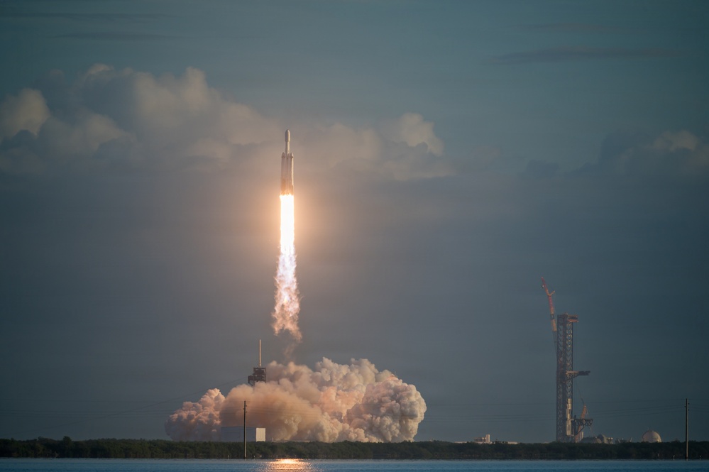 A large rocket is captured mid-launch from a coastal launch pad, producing a massive plume of white smoke and bright exhaust flame. The rocket's configuration — with a wide base suggesting multiple booster cores — is consistent with a heavy-lift vehicle. The scene is set against a partly cloudy sky with a body of water in the foreground and additional launch infrastructure visible to the right.