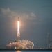 A large rocket is captured mid-launch from a coastal launch pad, producing a massive plume of white smoke and bright exhaust flame. The rocket's configuration — with a wide base suggesting multiple booster cores — is consistent with a heavy-lift vehicle. The scene is set against a partly cloudy sky with a body of water in the foreground and additional launch infrastructure visible to the right.