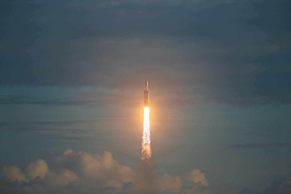 A large rocket is captured mid-ascent against an overcast, blue-grey sky. Intense engine exhaust produces a bright, glowing plume of fire and white smoke trailing below the vehicle. The rocket appears to have a multi-core or wide-body configuration visible at the top of the frame.