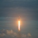 A large rocket is captured mid-ascent against an overcast, blue-grey sky. Intense engine exhaust produces a bright, glowing plume of fire and white smoke trailing below the vehicle. The rocket appears to have a multi-core or wide-body configuration visible at the top of the frame.