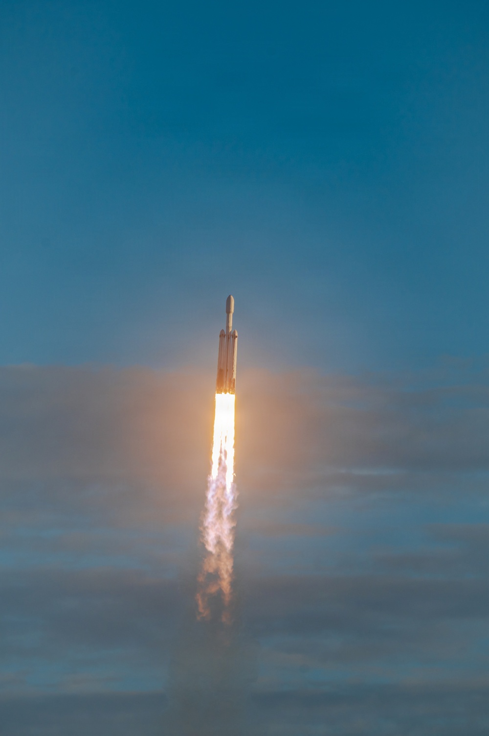 A large rocket is captured mid-flight ascending through a partly cloudy blue sky, with intense engine flames and a white exhaust plume trailing below it. The rocket's distinctive triple-core configuration is visible, consistent with a heavy-lift launch vehicle design. The image was taken from a distance, showing the rocket already well above the cloud layer.