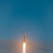 A large rocket is captured mid-flight ascending through a partly cloudy blue sky, with intense engine flames and a white exhaust plume trailing below it. The rocket's distinctive triple-core configuration is visible, consistent with a heavy-lift launch vehicle design. The image was taken from a distance, showing the rocket already well above the cloud layer.