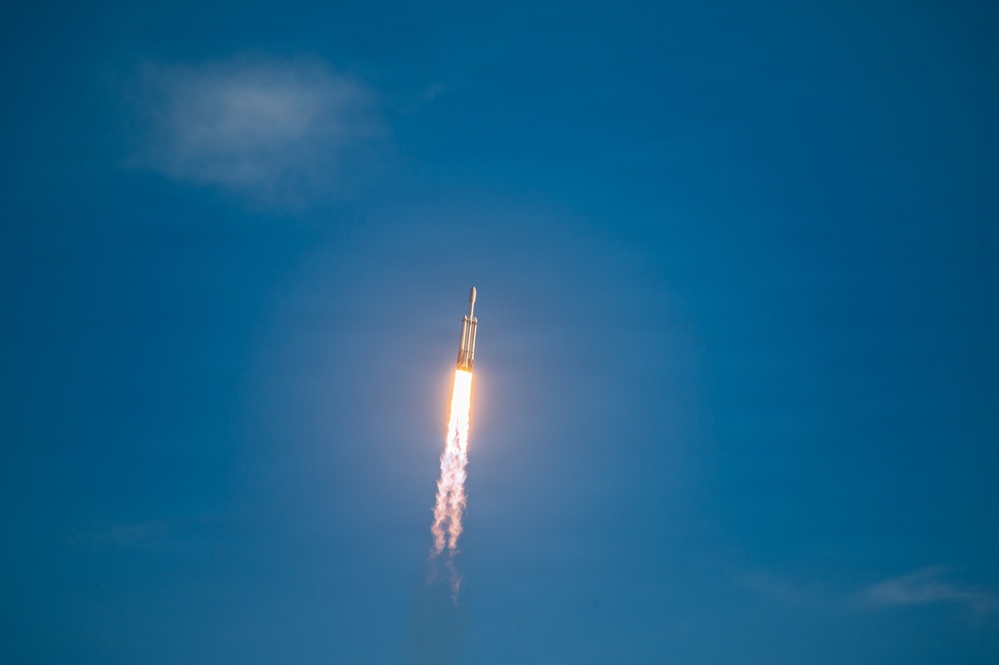 A rocket is captured mid-flight ascending through a clear blue sky, with a bright orange and white exhaust plume trailing below it. The rocket appears to be in its early ascent phase, climbing steeply upward. A single white cloud is visible in the upper left portion of the frame.