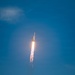 A rocket is captured mid-flight ascending through a clear blue sky, with a bright orange and white exhaust plume trailing below it. The rocket appears to be in its early ascent phase, climbing steeply upward. A single white cloud is visible in the upper left portion of the frame.