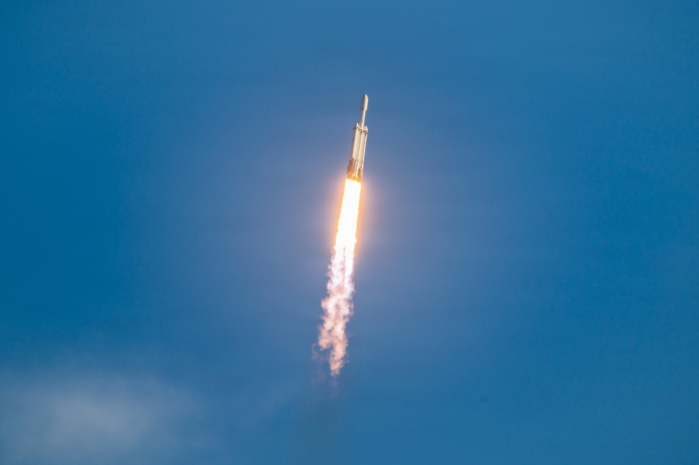 A large rocket is captured mid-ascent against a clear blue sky, producing a bright orange-white flame and a dense white exhaust plume trailing below it. The rocket's configuration — with a wide lower stage and a narrower upper stage — is consistent with a heavy-lift launch vehicle. The image was taken from a ground-level vantage point looking upward, capturing the rocket at an angle as it climbs steeply.