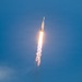 A large rocket is captured mid-ascent against a clear blue sky, producing a bright orange-white flame and a dense white exhaust plume trailing below it. The rocket's configuration — with a wide lower stage and a narrower upper stage — is consistent with a heavy-lift launch vehicle. The image was taken from a ground-level vantage point looking upward, capturing the rocket at an angle as it climbs steeply.
