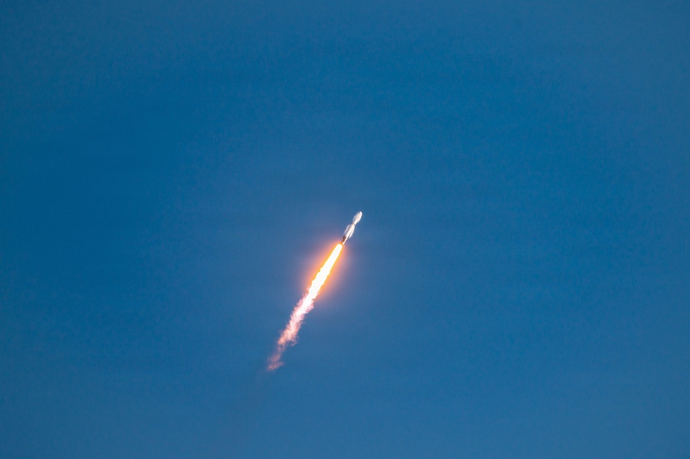 A rocket is captured mid-flight ascending through a clear blue sky at a steep diagonal angle. The rocket's engines are producing a bright orange-yellow flame and a white exhaust plume trailing behind it. The image appears to be taken from a ground-based vantage point during daytime conditions.