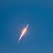 A rocket is captured mid-flight ascending through a clear blue sky at a steep diagonal angle. The rocket's engines are producing a bright orange-yellow flame and a white exhaust plume trailing behind it. The image appears to be taken from a ground-based vantage point during daytime conditions.