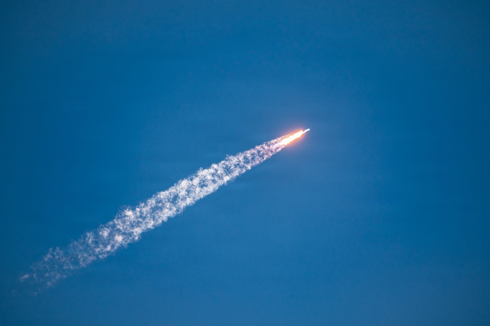 A rocket is captured mid-flight ascending diagonally through a clear blue sky. A bright orange-white flame is visible at the rocket's base, with a long white exhaust plume trailing behind it. The image appears to be taken from a ground-level vantage point using a telephoto lens.