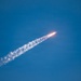 A rocket is captured mid-flight ascending diagonally through a clear blue sky. A bright orange-white flame is visible at the rocket's base, with a long white exhaust plume trailing behind it. The image appears to be taken from a ground-level vantage point using a telephoto lens.