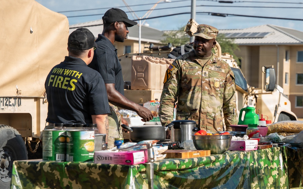 A U.S. Army soldier named Patterson, identifiable by his name tape and rank insignia on his OCP camouflage uniform, interacts with two civilians at an outdoor cooking or food preparation station. The table is covered with a camouflage cloth and holds various cooking equipment, canned goods, and food items. A military vehicle (likely an MRAP or similar tactical vehicle) is visible in the background, suggesting this is taking place on or near a military installation.