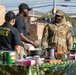 A U.S. Army soldier named Patterson, identifiable by his name tape and rank insignia on his OCP camouflage uniform, interacts with two civilians at an outdoor cooking or food preparation station. The table is covered with a camouflage cloth and holds various cooking equipment, canned goods, and food items. A military vehicle (likely an MRAP or similar tactical vehicle) is visible in the background, suggesting this is taking place on or near a military installation.