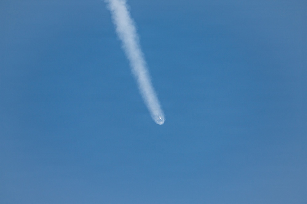 The image shows an aircraft or flying object at high altitude leaving a prominent white contrail (condensation trail) against a clear blue sky. The trail extends diagonally from the upper-left to the lower-right of the frame, with the object visible at the leading end. The sky is largely clear with minimal cloud cover.