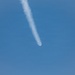 The image shows an aircraft or flying object at high altitude leaving a prominent white contrail (condensation trail) against a clear blue sky. The trail extends diagonally from the upper-left to the lower-right of the frame, with the object visible at the leading end. The sky is largely clear with minimal cloud cover.