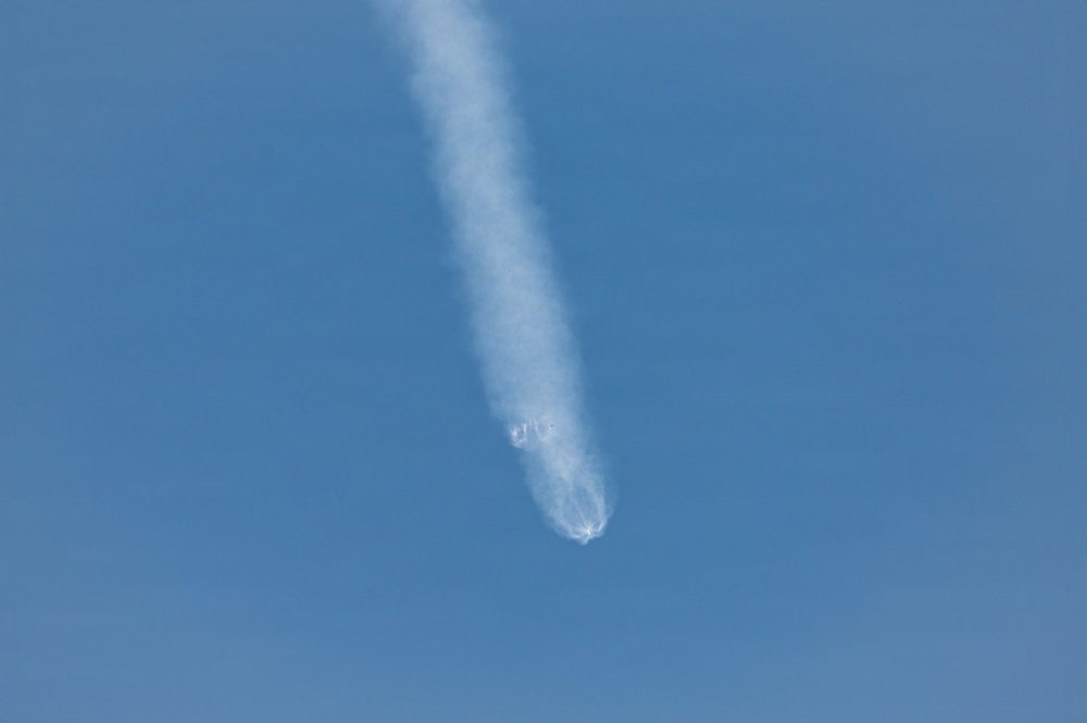 The image shows an aerial object trailing a white smoke/vapor plume against a clear blue sky. At the lower end of the trail, there appears to be a bright flash or explosion with debris dispersing outward, suggesting a mid-air breakup or detonation event. The long, straight contrail above indicates the object had been traveling at high altitude before the apparent failure.