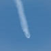 The image shows an aerial object trailing a white smoke/vapor plume against a clear blue sky. At the lower end of the trail, there appears to be a bright flash or explosion with debris dispersing outward, suggesting a mid-air breakup or detonation event. The long, straight contrail above indicates the object had been traveling at high altitude before the apparent failure.