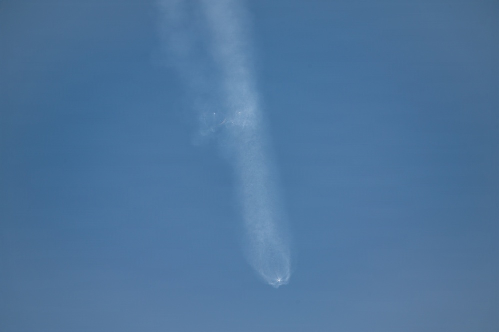 The image shows a rocket or missile in flight against a clear blue sky, leaving a visible exhaust plume or contrail. The trail appears to widen toward the bottom of the frame, suggesting the object is traveling upward. A bright point of light or small explosion-like feature is visible near the middle of the trail.