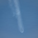 The image shows a rocket or missile in flight against a clear blue sky, leaving a visible exhaust plume or contrail. The trail appears to widen toward the bottom of the frame, suggesting the object is traveling upward. A bright point of light or small explosion-like feature is visible near the middle of the trail.