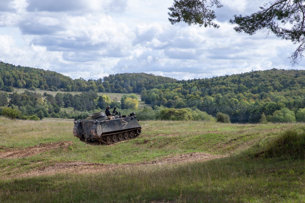 A tracked armored personnel carrier (APC), consistent with an M113 variant, is traversing an open grassy field during what appears to be a military exercise or training operation. At least one crew member is visible in the open hatch at the top of the vehicle. The setting is a rolling, wooded landscape under a partly cloudy sky.