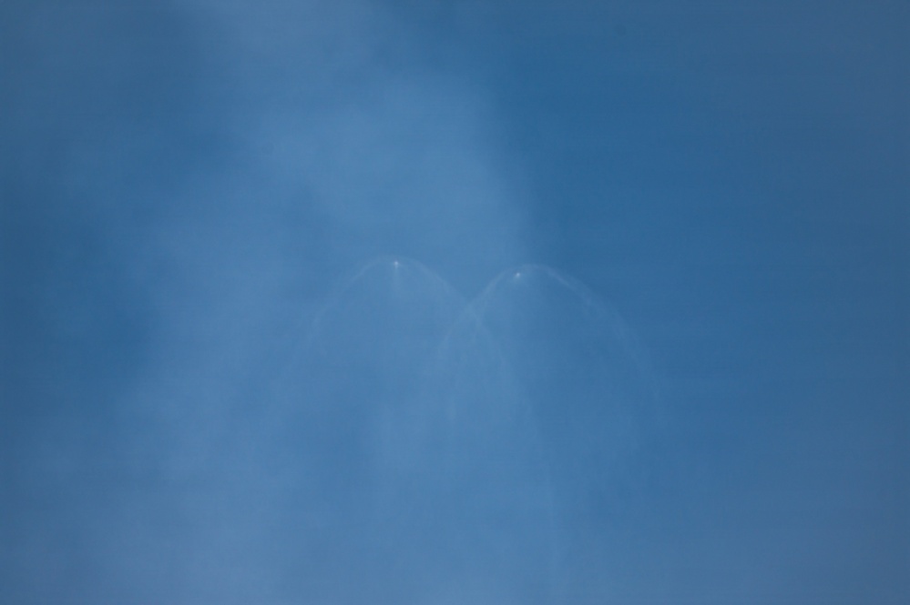 The image shows a blue sky with thin, wispy cloud formations or contrails forming a V-shape or wing-like pattern near the center of the frame. Two small bright points are visible at the apex of the V-shape, possibly aircraft or light reflections. The overall image is hazy and low in contrast.